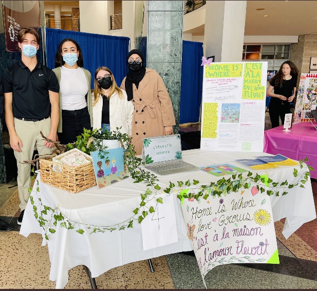 Ella, Noah, Raya, and Federica standing behind their project presentation booth at Youth Action Showcase. The table has a white tablecloth, green vines loosely wrapped around it, and a poster board on the right side.