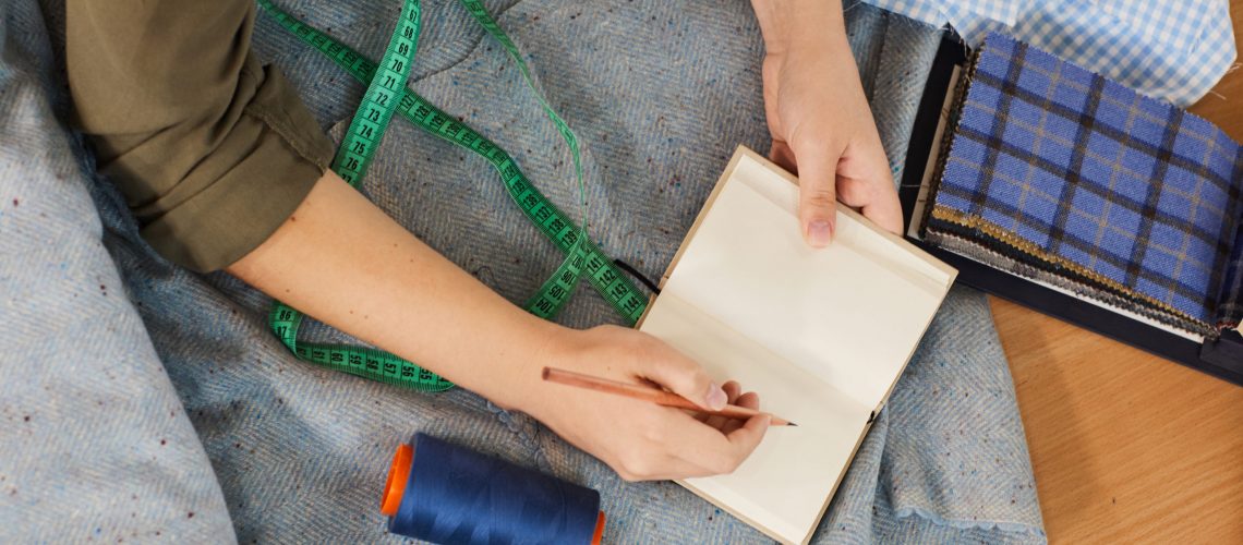 Student learning to sew Close-up of female student opening her notepad and making notes during sewing lesson at the table in workshop